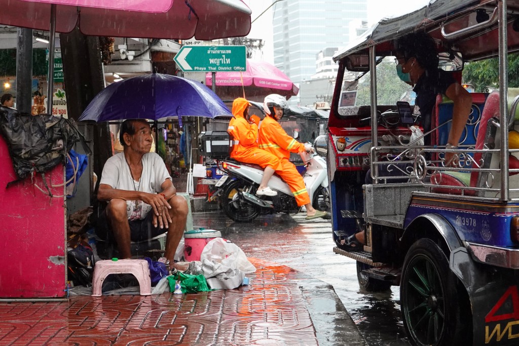Photo by CHAYANIT ITTHIPONGMAETEE / AFP A street vendor sits under an umbrella to shield from the rain as he talks to a tuk-tuk driver while waiting for customers in Bangkok on September 26, 2026.