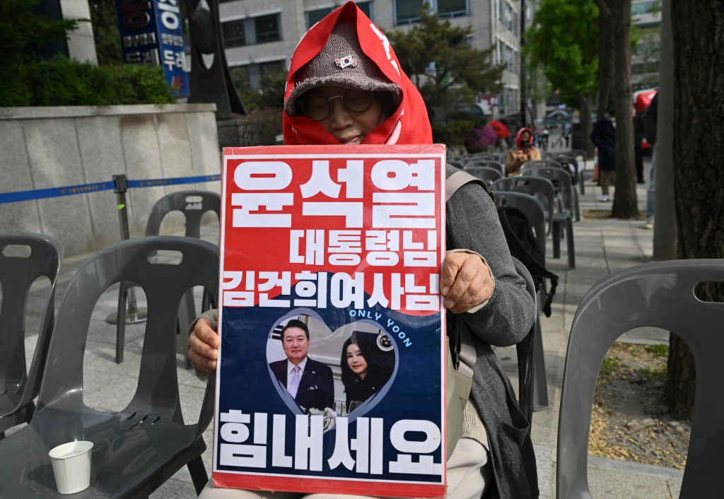 Photo by JUNG YEON-JE / AFP  A supporter of South Korea's former president Yoon Suk Yeol and his wife Kim Keon Hee holds a placard showing a picture of Yoon and Kim as she watches a live stream of the trial of the former first lady on a street near the Seoul High Court in Seoul on April 28, 2026.