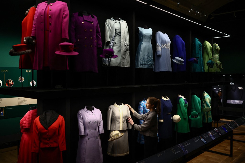 Photo by HENRY NICHOLLS / AFP  A member of the Royal Collection Trust staff (C) poses next to a suit 'Silk Starron', circa 1960, during a media preview of the exhibition 'Queen Elizabeth II: Her Life in Style' at the King’s Gallery in Buckingham Palace in London on April 9, 2026.