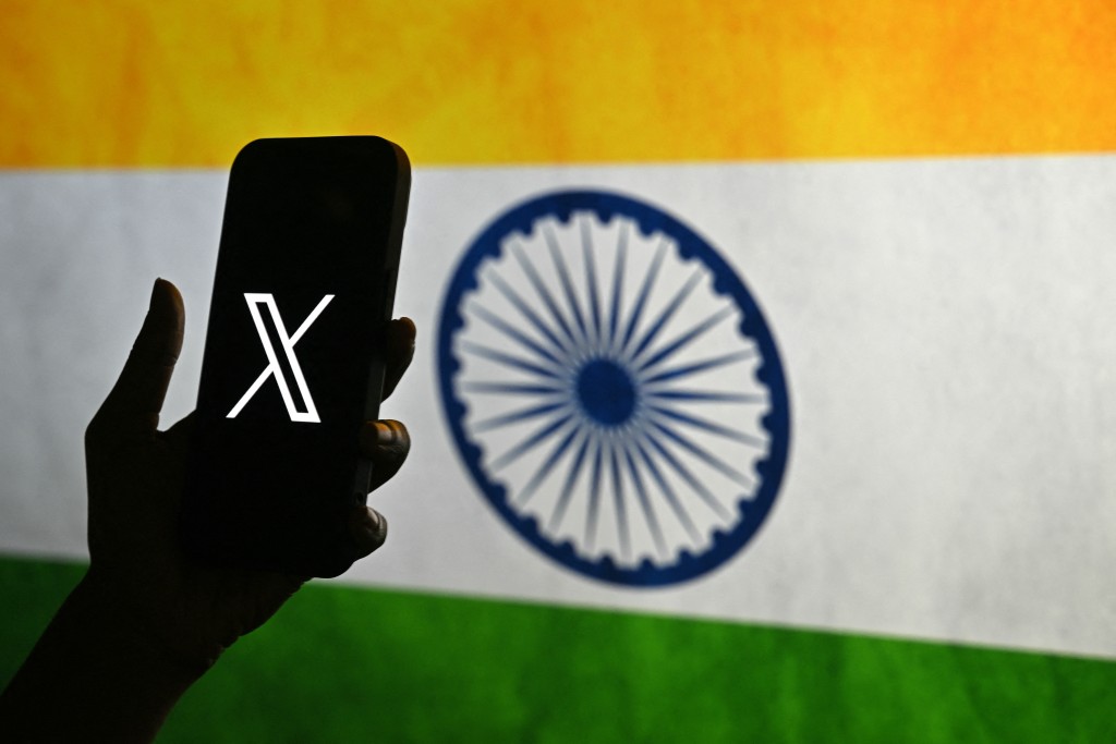 Photo by IDREES MOHAMMED / AFP. This photo illustration shows a person holding a smartphone bearing the logo of X, formerly known as Twitter, in front of the Indian national flag in Bengaluru on January 15, 2026.